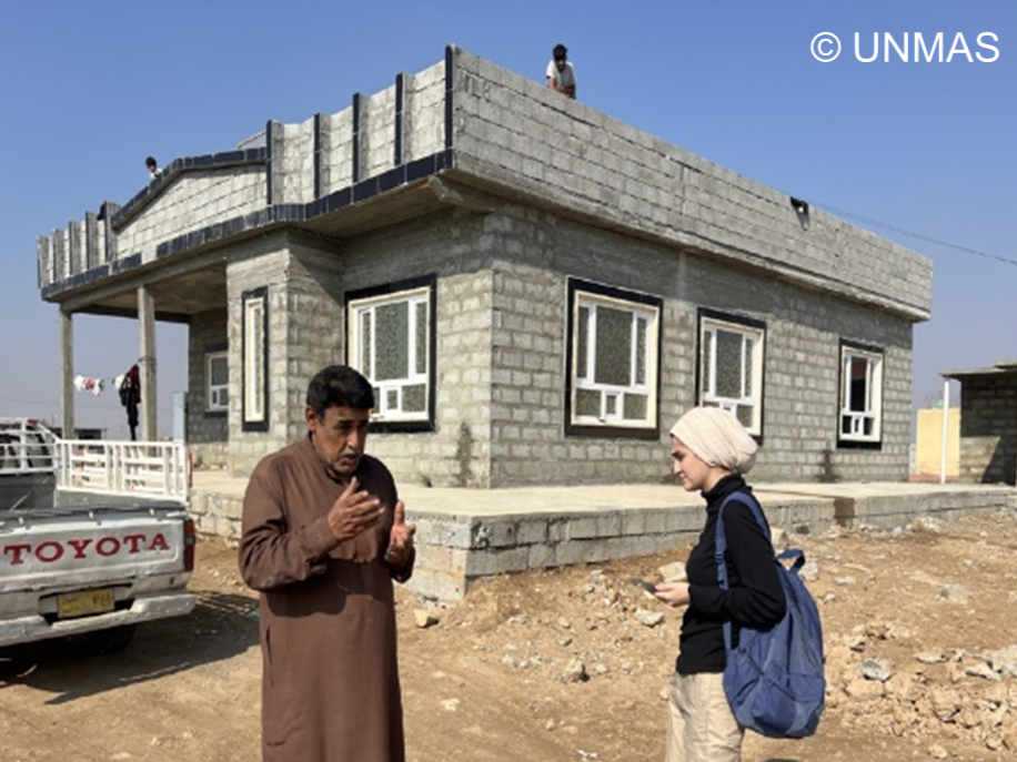 A man with brown robe is talking with a woman with black shirt in front of a newly constructed house.