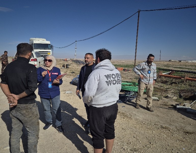 A woman wearing UNMAS jacket is talking to a group of men on a work site.