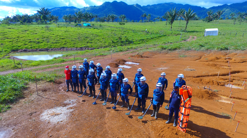 A group of people in blue uniforms and protective helmets stand in a line on a dirt field, each holding a metal detector.