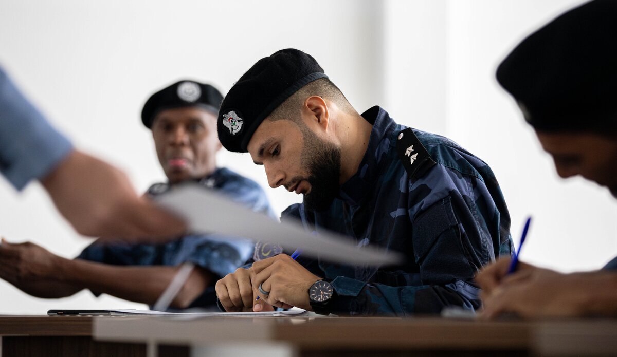 A group of uniformed individuals, likely military personnel, seated at desks in a classroom setting. The central figure, wearing a dark beret with an emblem and a camouflage uniform, is writing on paper with a pen. Another similarly dressed individual is visible in the background, also engaged in writing or reading. The scene conveys a training or educational environment.