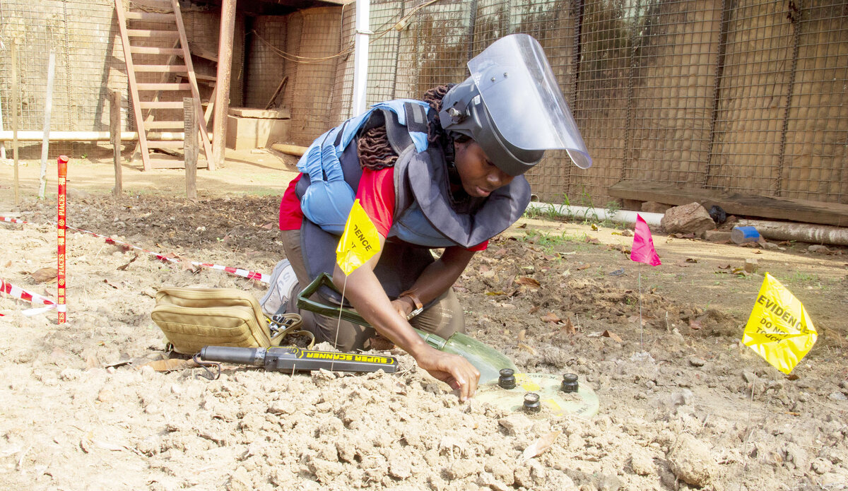 A deminer wearing protective gear and a face shield carefully examines the ground with demining tools and evidence flags during a mine clearance operation.