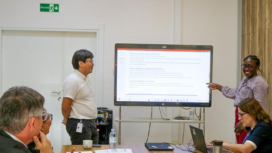 A group of people in a meeting room are gathered around a large digital screen displaying text. One person stands and points at the screen, while another stands nearby. Two others are seated at the table with laptops and documents in front of them. The room has a white wall with a green exit sign and a wooden panel accent.