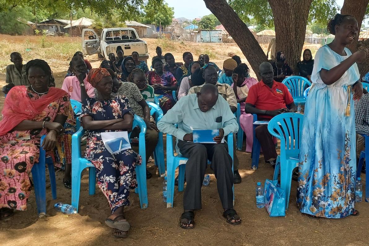 When given an opportunity to ask UNMISS and RJMEC representatives about the political situation in South Sudan, community members in Tokiman East near Juba grabbed it with both hands. Photos: James Ohisa/UNMISS A group of people sitting in blue plastic chairs underneath a tree. In the background, there is a car with its door open.