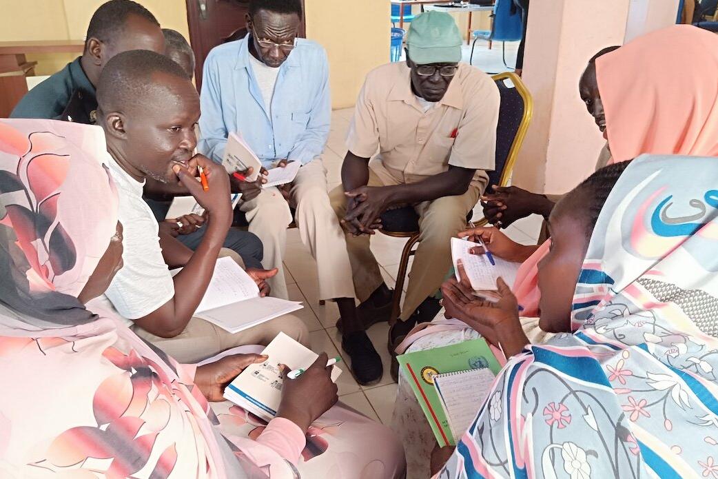 A group of people standing outdoors on a grassy field, some holding hands and others raising arms, appearing to participate in a group activity or celebration. Footwear is scattered on the ground, and a partly cloudy sky is visible in the background.
