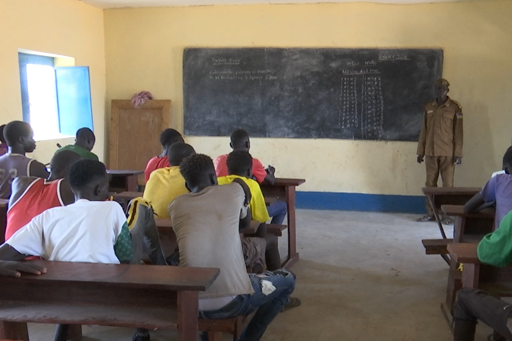 Classroom scene with students seated at wooden desks facing a chalkboard, where mathematical calculations and notes are written. An instructor stands near the board in front of the class.