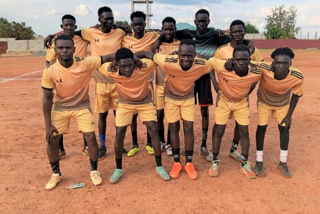 A soccer team wearing matching yellow and black uniforms poses together on a dirt field, with goalposts and trees visible in the background