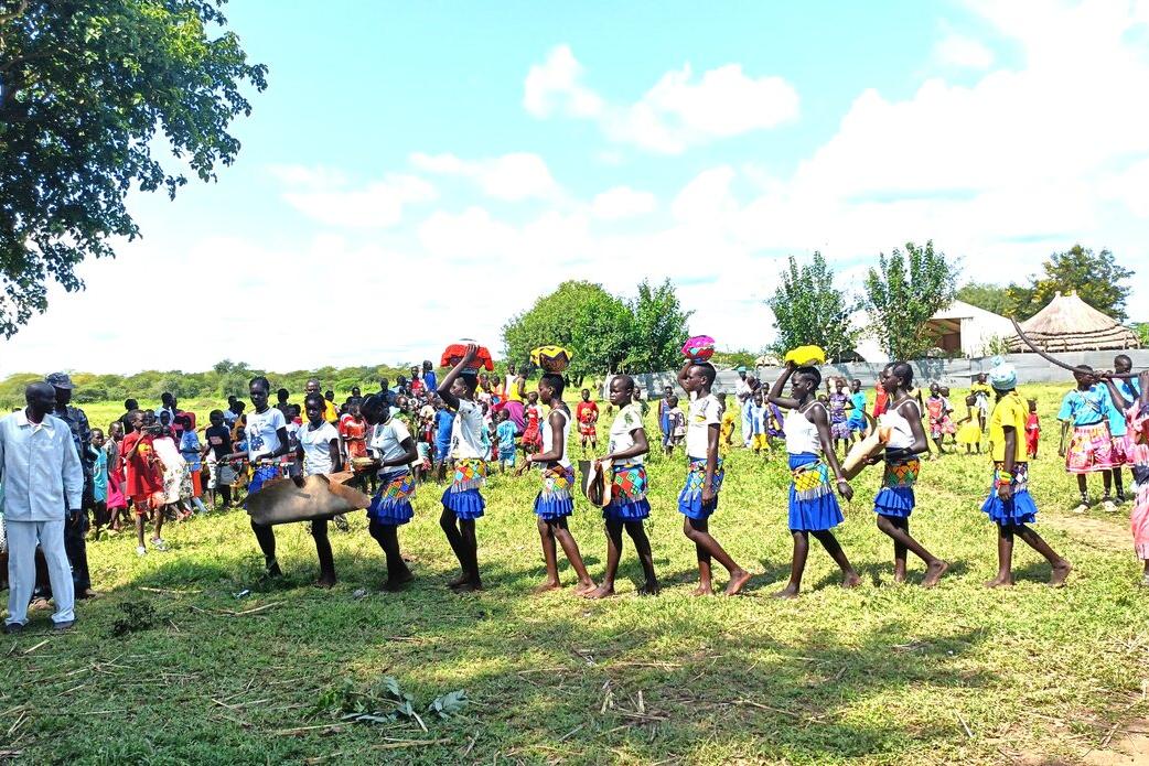 A group of children in colorful clothing and blue skirts perform a traditional dance in a grassy field, with a crowd of spectators and trees in the background.