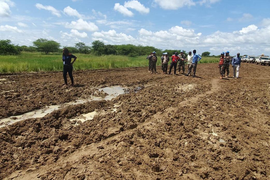 A group of people, including uniformed personnel, stand on a muddy road with puddles, near grassy fields and parked vehicles under a partly cloudy sky.