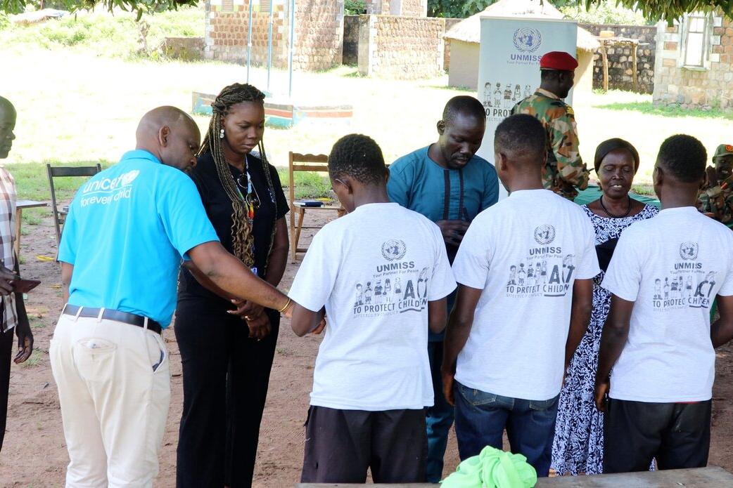 Three children stand under mango trees with a few belongings as they are released from South Sudanese armed forces and begin a new life. Group of people standing outdoors in a circle, including individuals wearing UNMISS-branded shirts and a person in a UNICEF shirt, with soldiers and chairs in the background.