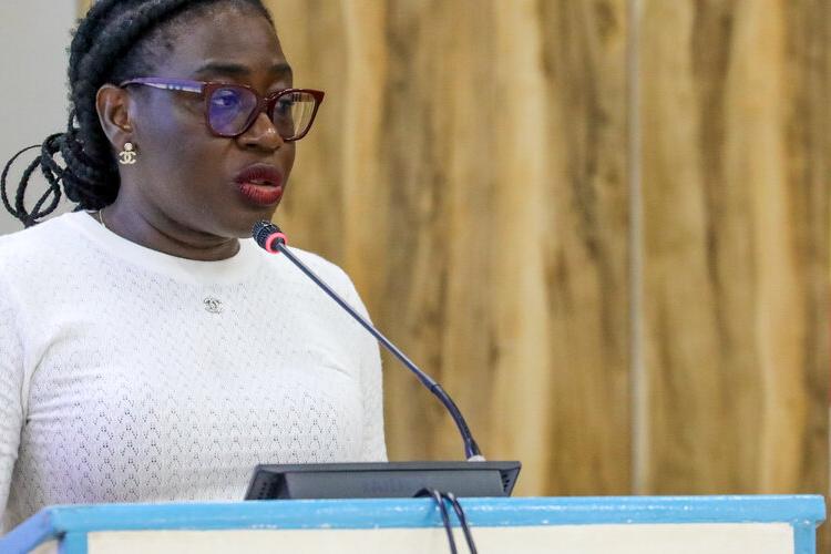 A young woman standing at a podium speaking into a microphone with a flag in the background.