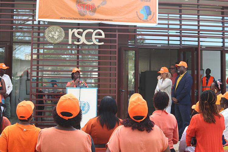 The backs of people all wearing orange facing a stage where a young woman in an orange baseball cap is addressing the crowd.