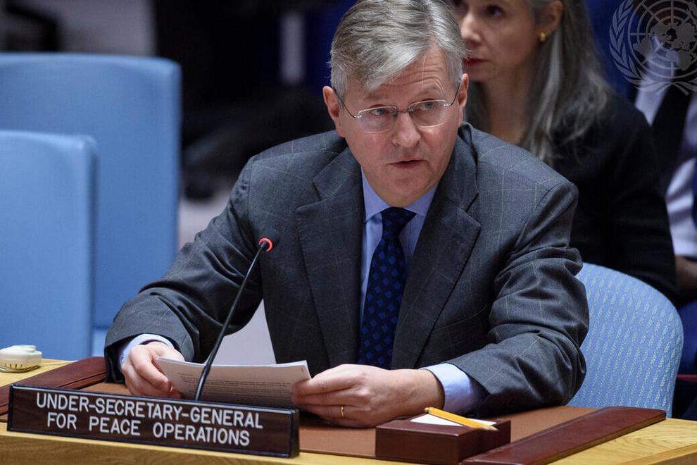 A man in a grey suit sitting a desk behind a sign indicating he is the Under-Secretary-General for Peace Operations. He is speaking into a microphone and addressing the room.