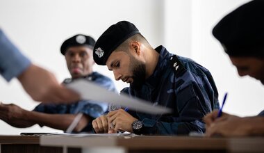 A group of uniformed individuals, likely military personnel, seated at desks in a classroom setting. The central figure, wearing a dark beret with an emblem and a camouflage uniform, is writing on paper with a pen. Another similarly dressed individual is visible in the background, also engaged in writing or reading. The scene conveys a training or educational environment.