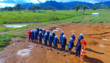 A group of people in blue uniforms and protective helmets stand in a line on a dirt field, each holding a metal detector. One individual in red stands separately. The background includes green fields, palm trees, mountains, a small pond, and dug-out areas under a partly cloudy blue sky.