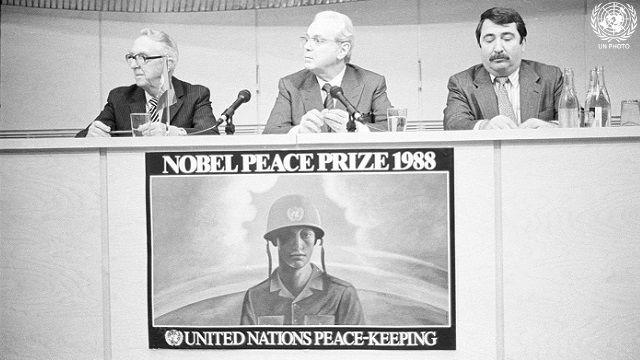 A black-and-white photo of three men behind a podium, above a poster that reads Nobel Peace Prize 1988, United Nations Peacekeeping